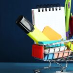 A mini shopping cart is holding a notebook, pens, highlighter and other stationery supplies. Part of an article: Build the Perfect Bujo with these Bullet Journal Supplies for Beginners