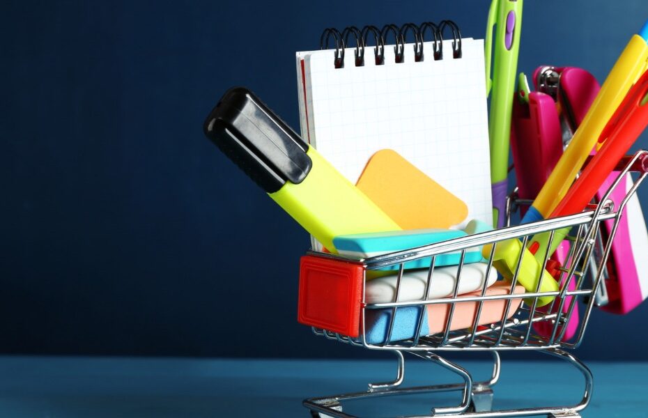 A mini shopping cart is holding a notebook, pens, highlighter and other stationery supplies. Part of an article: Build the Perfect Bujo with these Bullet Journal Supplies for Beginners