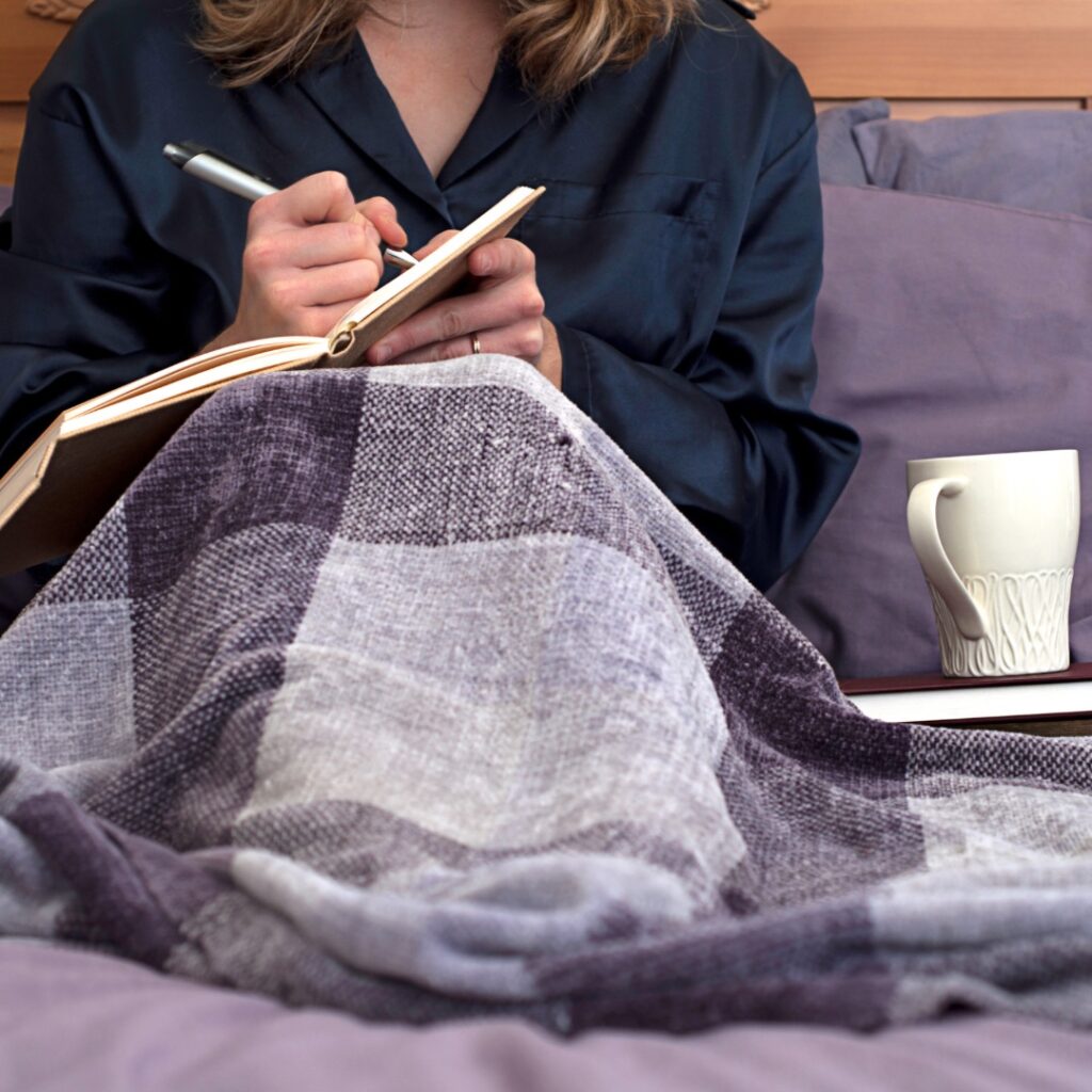 A woman sits up in bed. She has a blanket over her, we don’t see her face. She has a notebook open and is writing in it. She has more books at her side with a mug resting on the top.