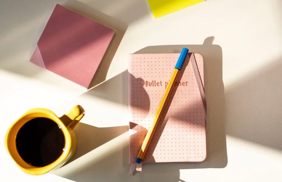 A bullet journal, pen , memo block and cup lay flat on a table. Part of an article about what to include in a bullet journal